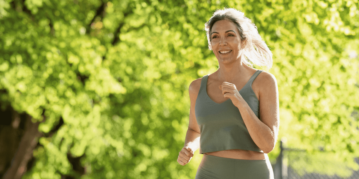 A smiling woman jogging outdoors against a background of green leaves.